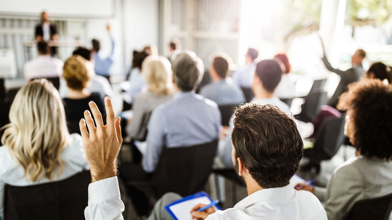 Rear view of a businessman raising his hand to ask the question on a seminar with large group of his colleagues.